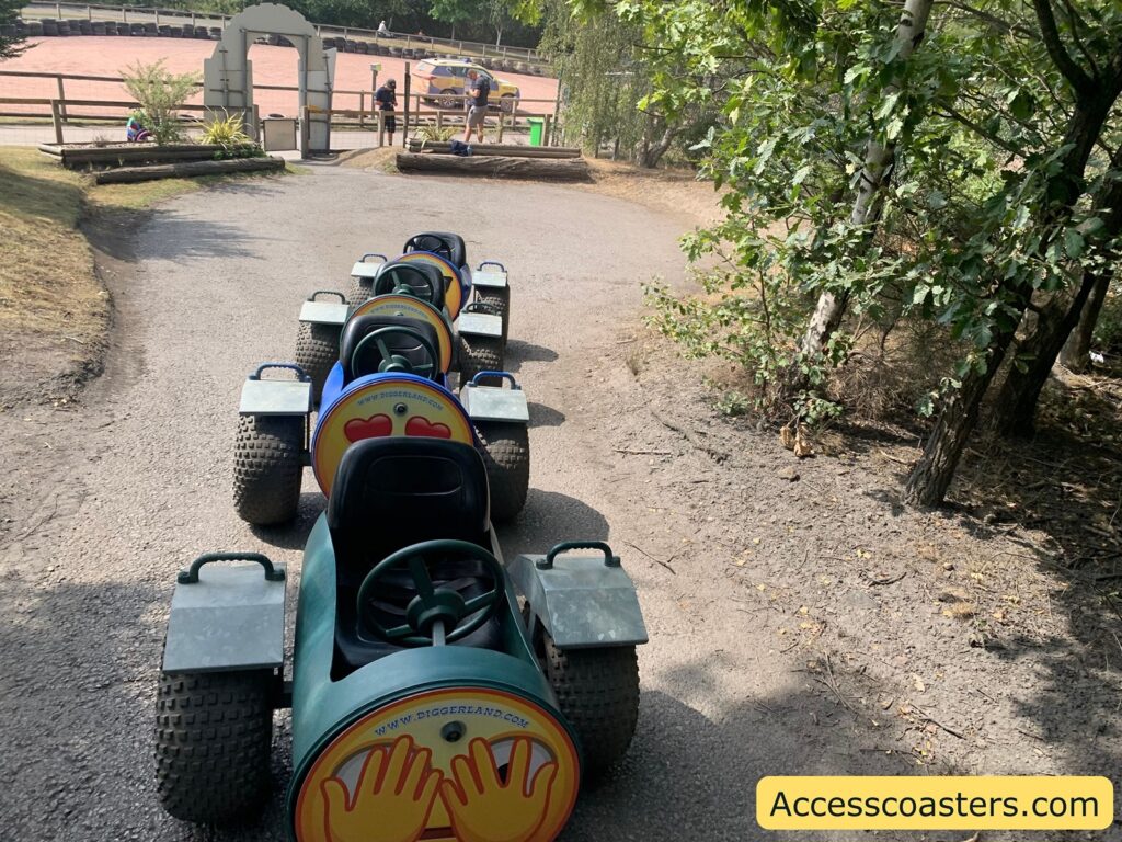 A row of four small, off-road style go-karts with wide tires lined up on a paved path. The backs of the karts display circular signs with two yellow hands covering a red heart,