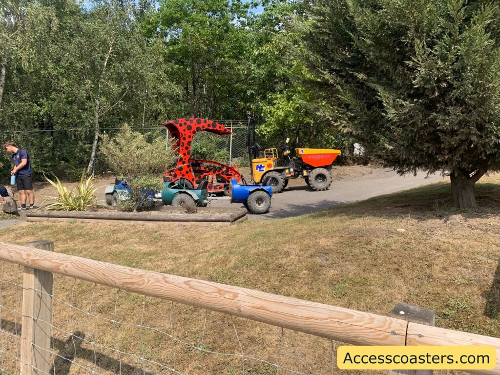 train made up of a digger at the front with ride viclas behind. the front carrage of the train is is painted with red and black spots, reminiscent of a ladybird pattern, and has a whimsical, exaggerated roof. the behind carages are red and have emjo faces on teh front