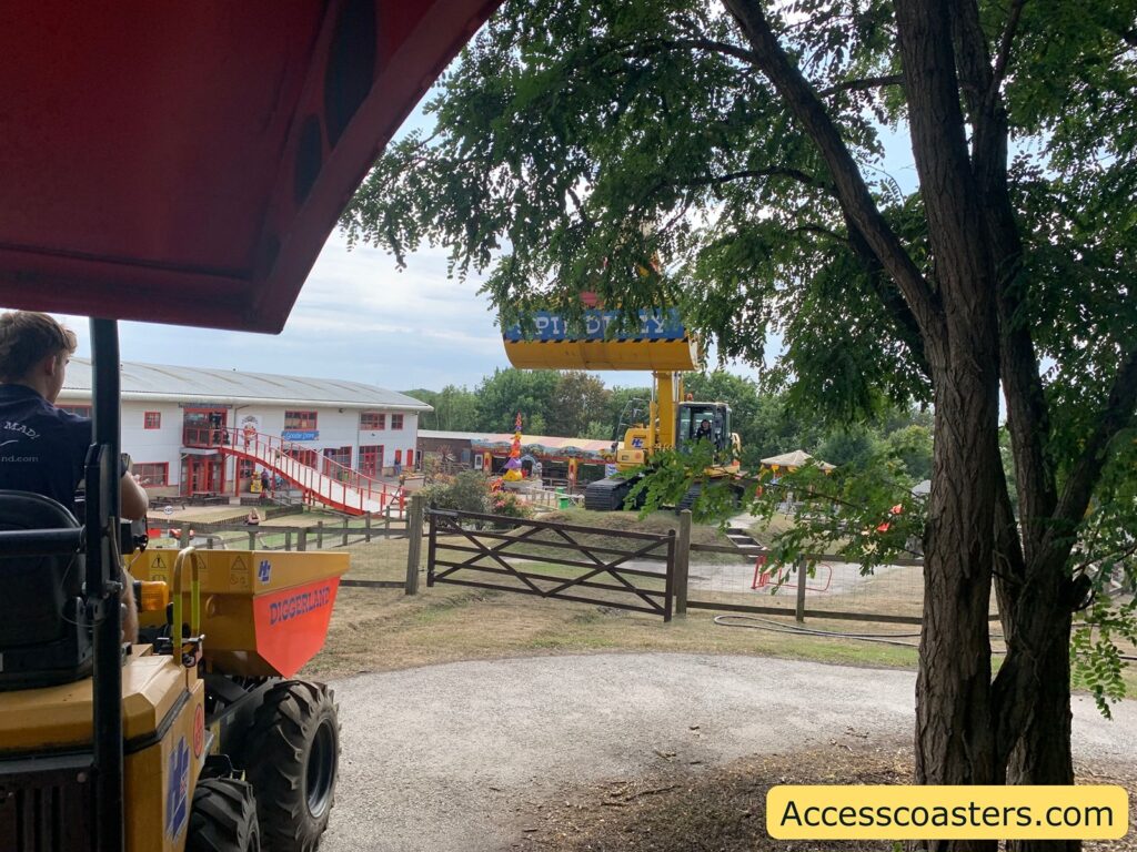 In the foreground, there is a person driving a yellow construction-themed ride vehicle labeled "DIGGERLAND" under a partially shaded area, with a tree to the right. In the middle distance, there is a fenced-off area with more construction-themed machinery and a large yellow ride structure To the left, you can see a white building with red trim and red accents, and an elevated walkway or slide leading to it.