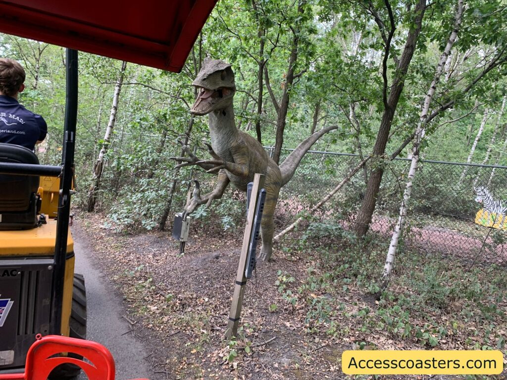 Animatronic dinosaur display in a wooded area, viewed from a moving ride vehicle. The dinosaur is positioned on the right side of a path, behind a fence and among trees