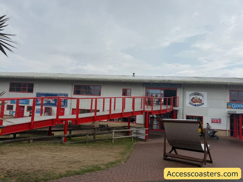 Outdoor scene showing the entrance to a leisure building with a wide red and white wheelchair-accessible ramp leading up to the second floor. The building has a light grey exterior, red window frames, and several signs, including one welcoming visitors, a "Kart World" logo, and signage for "The Dig Inn" upstairs.