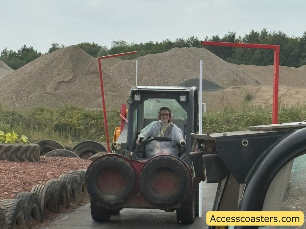 Iona driving a small construction loader vehicle outdoors, on a path lined with old tires.