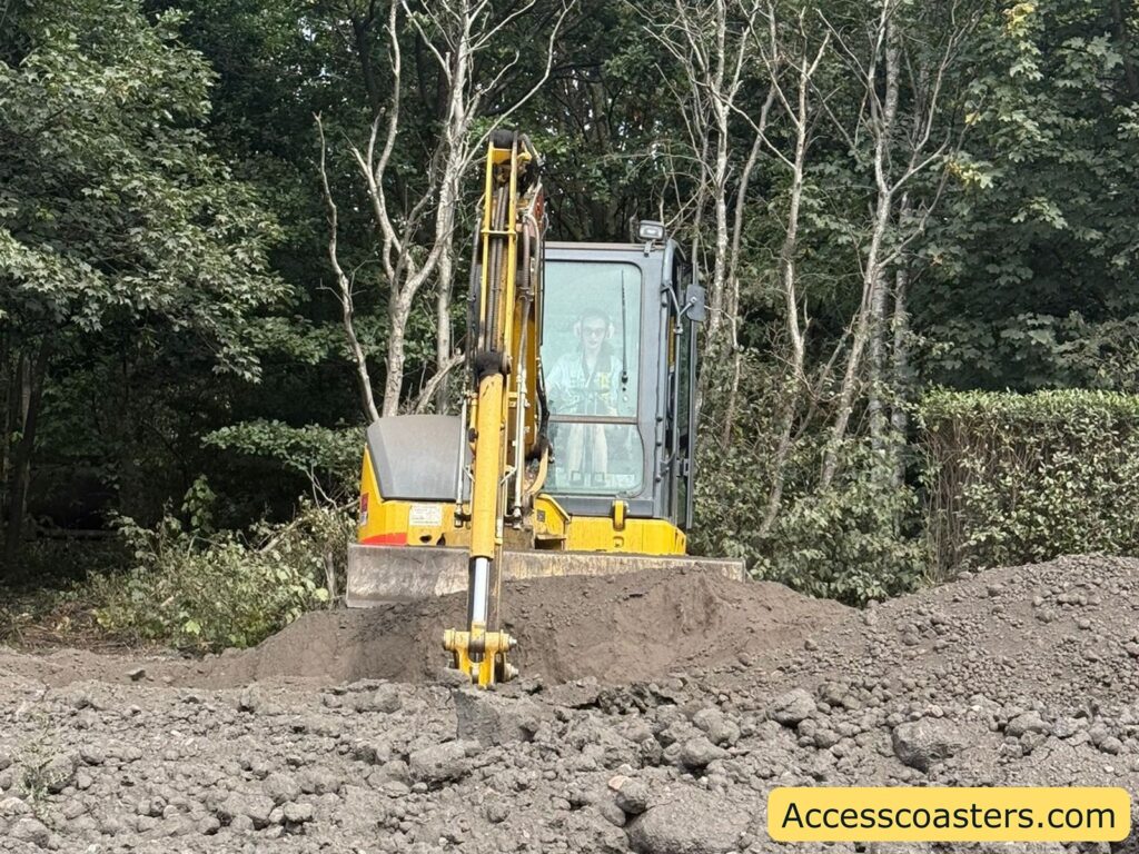 Excavator in a forested or wooded area, positioned on rough ground and dirt piles. The machine is yellow, and Iona is visible inside the cab,