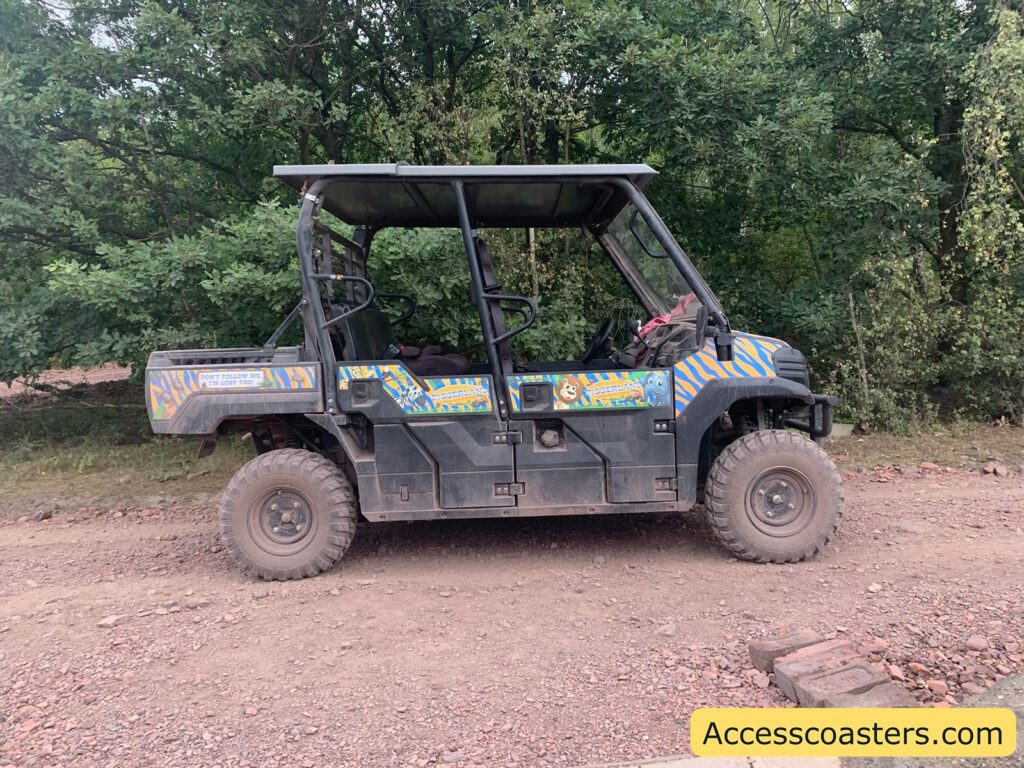 Utility terrain vehicle (UTV) with off-road tires parked on a dirt path beside a wooded area. The vehicle has colorful blue, orange, and yellow striped graphics on its sides with cartoon animal characters and the word "DIGGERLAND." There is a sign on the rear side panel that says, "DON'T FOLLOW ME, I'M LOST TOO!" The vehicle has a roof and open sides, with seating for several people.