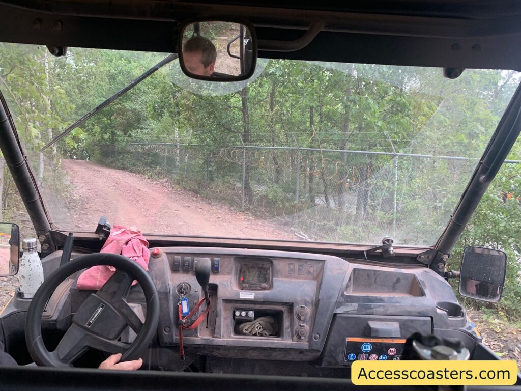 Interior view of an off-road utility vehicle, looking out through the windshield at a dirt path winding to the left. The dashboard is dusty, with a steering wheel on the left, various switches and keys, a gear shifter