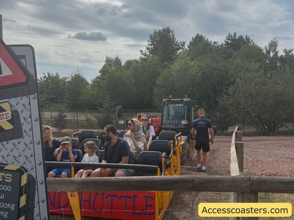 several people seated in a themed vehicle that is a digger. The ride is outdoors on a gravel path, with dense green trees and a cloudy sky