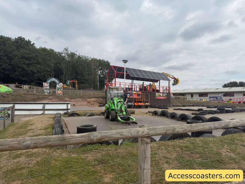 green tractor-like vehicle parked on a flat, square platform surrounded by large black tires, the arm of the tractor has one tire in the air