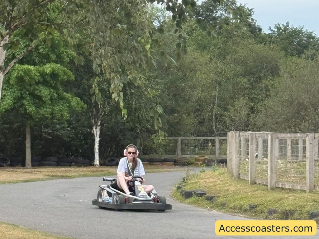 Go-kart track outdoors, surrounded by greenery and trees. ona's driving a go-kart, wearing ear defenders, sunglasses, and a lanyard. The track is bordered by black tires and a wire fence on one side.
