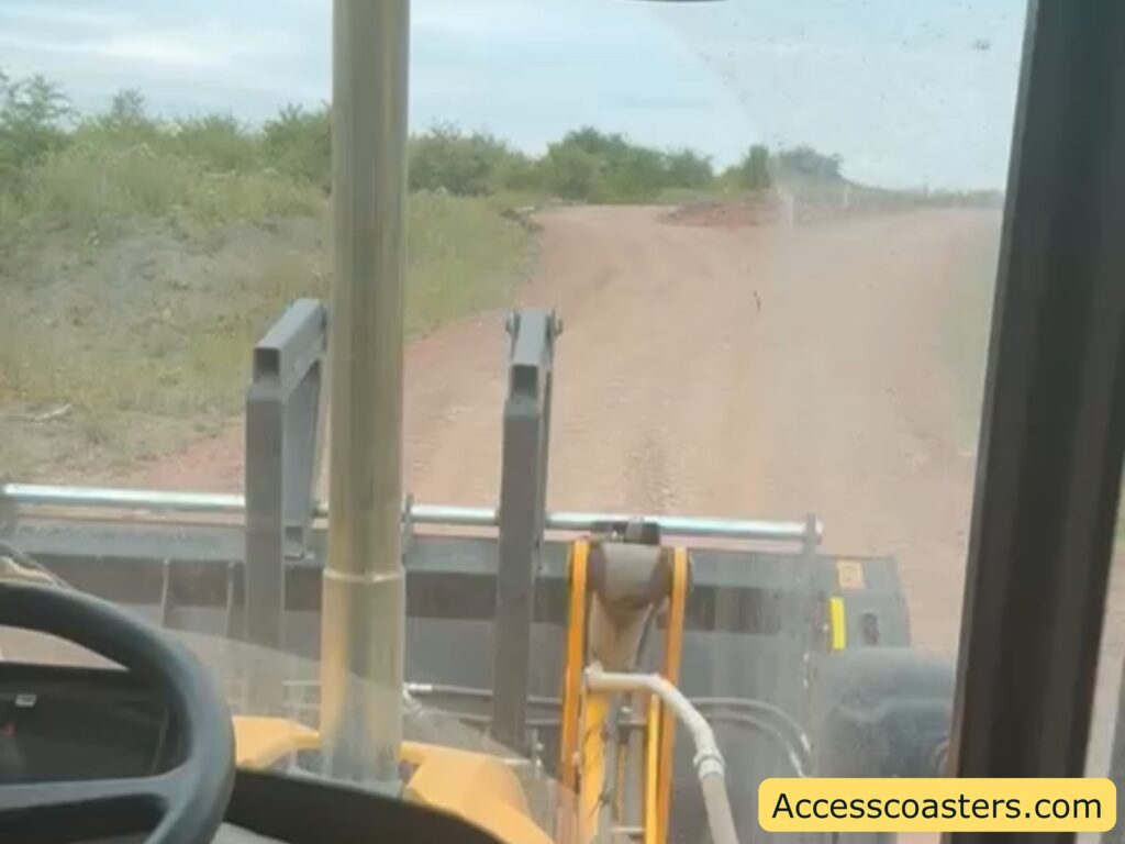 View from inside a construction vehicle, looking out onto a dirt road surrounded by grass and low shrubs. Ahead, the path curves slightly to the right. The vehicle's large implement is visible just outside the window.