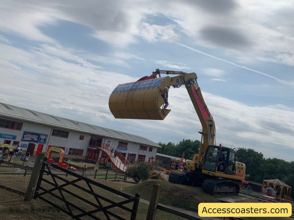 A large yellow excavator has been modified so its bucket holds a rotating ride vehicle labeled “SPINDIZZY.” The vehicle is shaped like a large drum or barrel, and passengers are strapped inside as the whole assembly is lifted and spun by the excavator’s arm. The ride is surrounded by a fence and a grassy area.