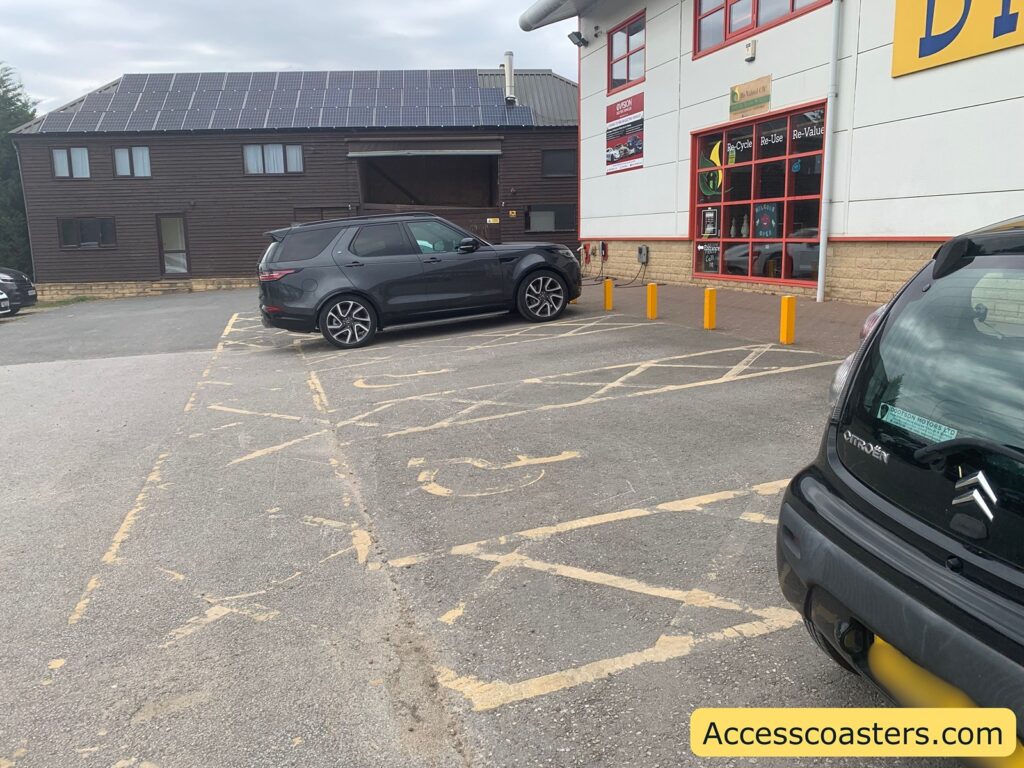 car park in front of a building with multiple vehicles. The area in the foreground and near the building features yellow-highlighted markings on the ground, indicating accessible or disabled parking spaces, although the lines are quite worn and faded
