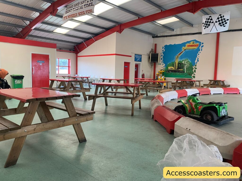 ndoor picnic area with wooden benches and tables, red and white decor, and a small children's go-kart track. In the foreground, there’s a green ride-on car behind plastic safety barriers. The walls are mostly white with red trim and feature a colorful mural with construction vehicles