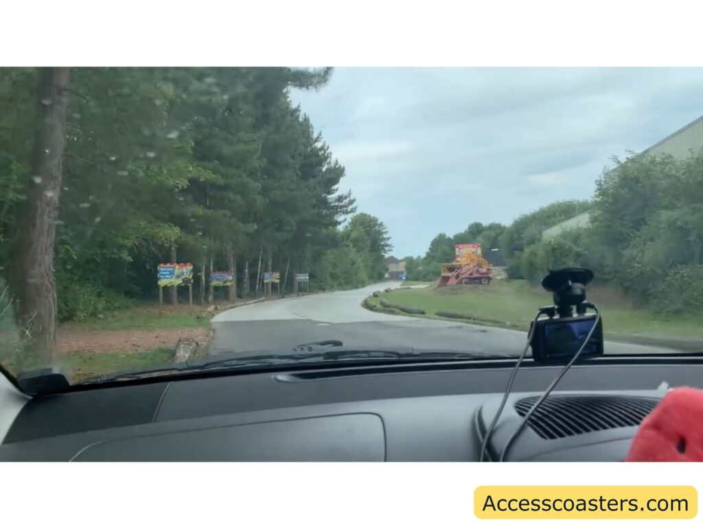 View from inside a car looking through the windshield at a curved road lined with trees on the left. Several colorful signs stand along the left sideall saying diggerland and directing to the car park