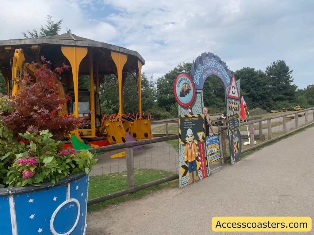 ride entrance themed around construction vehicles. The sign above the entrance arch reads "DIGA-ROUND Around We Go!" with additional signage featuring cartoon construction workers and ride restrictions, such as minimum height requirements. The attraction itself is visible on the left—a carousel-like ride with seats attached to what appears to be an actual digger or excavator machinery.