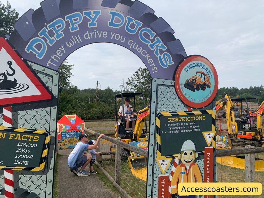 Colorful entrance sign at the "Dippy Ducks" attraction in Diggerland, an amusement park themed around construction equipment. The archway sign reads "DIPPY DUCKS - They will drive you quackers!" set on a background designed to look like a tire tread. On the left, information signs list fun facts about the ride, including costs and weights, and there is a playful warning sign with a cartoon duck in water. A larger sign on the right gives ride restrictions, which say the minimum height to ride is 80cm if accompanied by an adult and 110cm to operate. There is a cartoon construction worker in high-visibility clothing pointing to the height requirements.