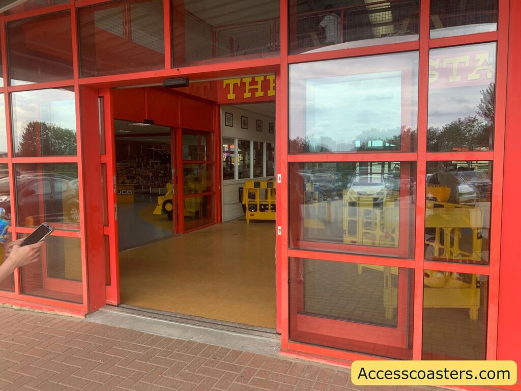 Entrance from the car park to the diggerland reception to a with large red-framed glass doors, partially open to reveal a spacious interior. Just inside the entrance, there are two yellow construction-themed rides for children.