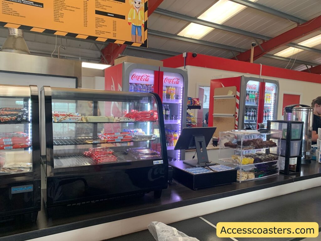 Café counter with snack display cases containing chocolate bars, cakes, and cookies. Behind the counter, there are refrigerated Coca-Cola branded fridges stocked with various soft drinks. Above the counter is a large orange menu board listing food and drink options with prices