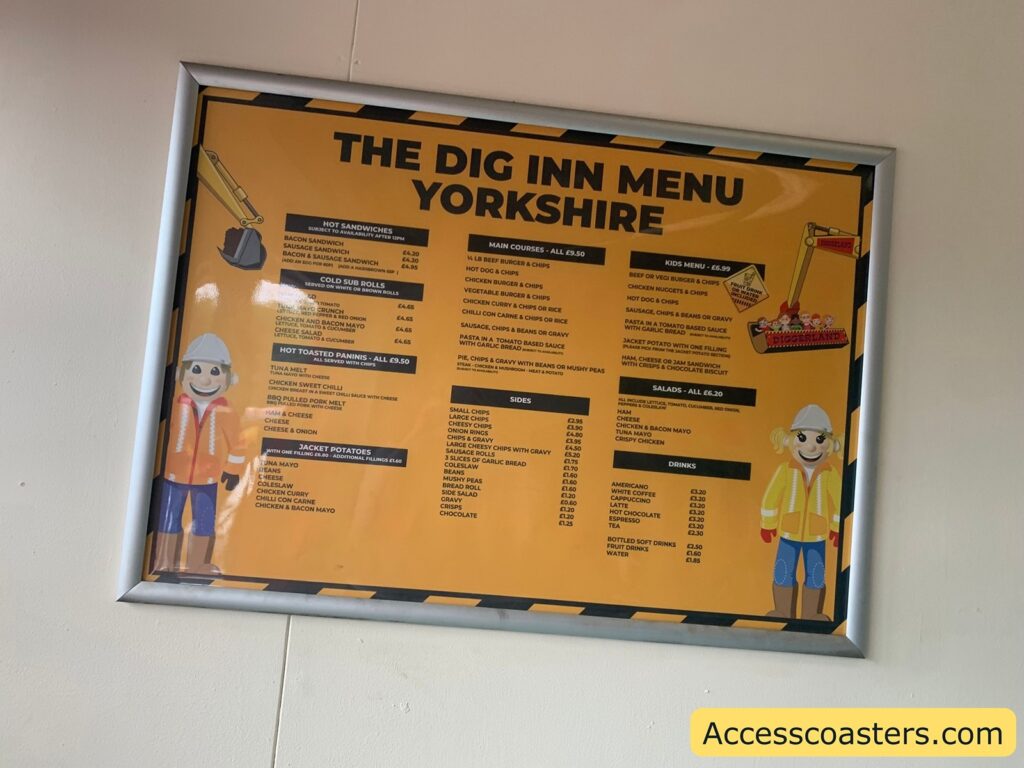 Framed restaurant menu on a yellow and black background, titled "The Dig Inn Menu Yorkshire." The menu features sections for hot sandwiches, cold sub rolls, hot toasted paninis, jacket potatoes, main courses, sides, kids menu, salads, and drinks. Cartoon construction workers, one on each side, wear orange safety jackets and white helmets.