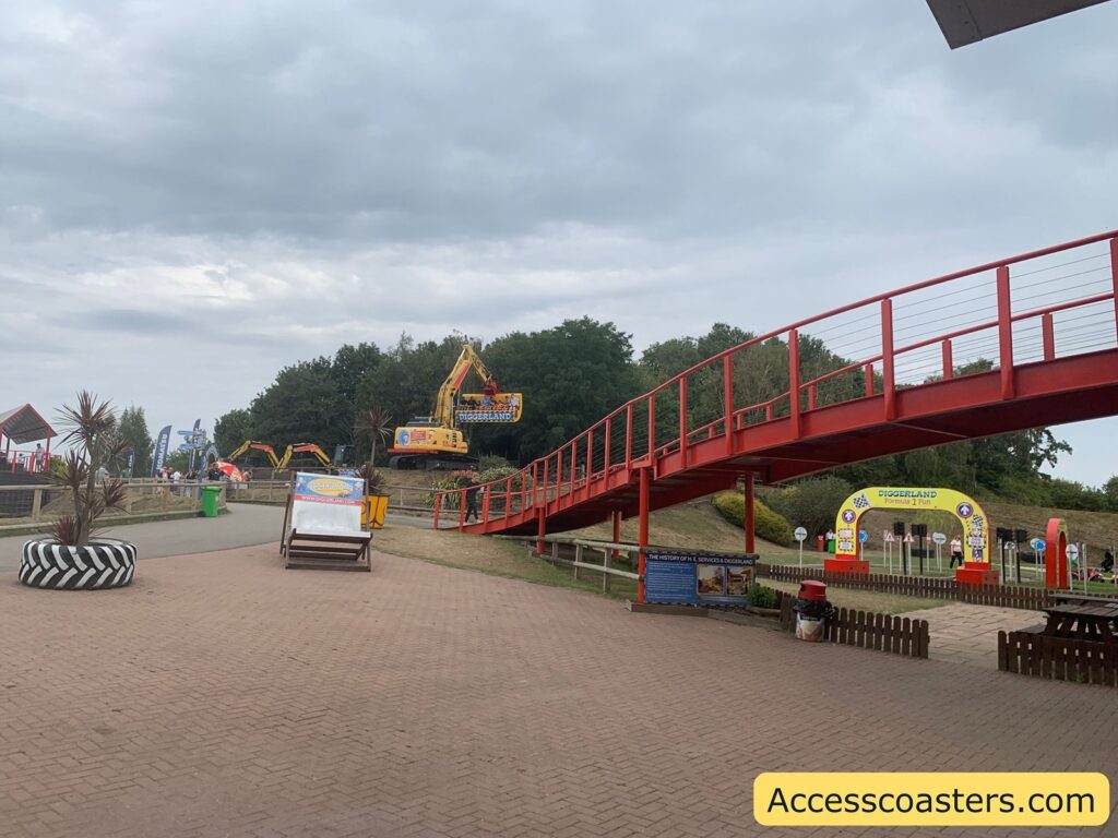 Outdoor amusement park scene featuring a paved walkway, a bright red pedestrian bridge crossing over it, and several construction-themed attractions visible in the background. Large yellow construction vehicles, such as excavators with "Diggerland" branding, are positioned as part of the attractions. There are informational signs, a large deckchair with promotional graphics, a circular planter made from a tire.