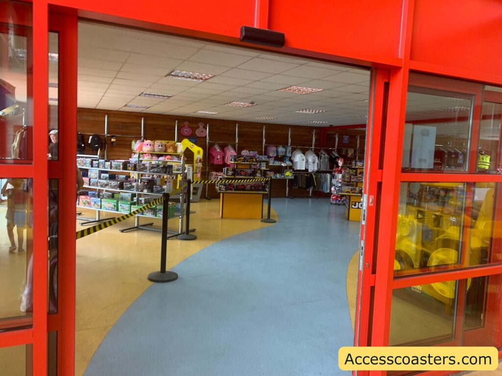 Gift shop entrance with red framed doors, black and yellow caution tape stretched across the entrance, and shelves displaying toys, merchandise, and various souvenir items inside. there is yellow barrer leading to a counter for the reseption area