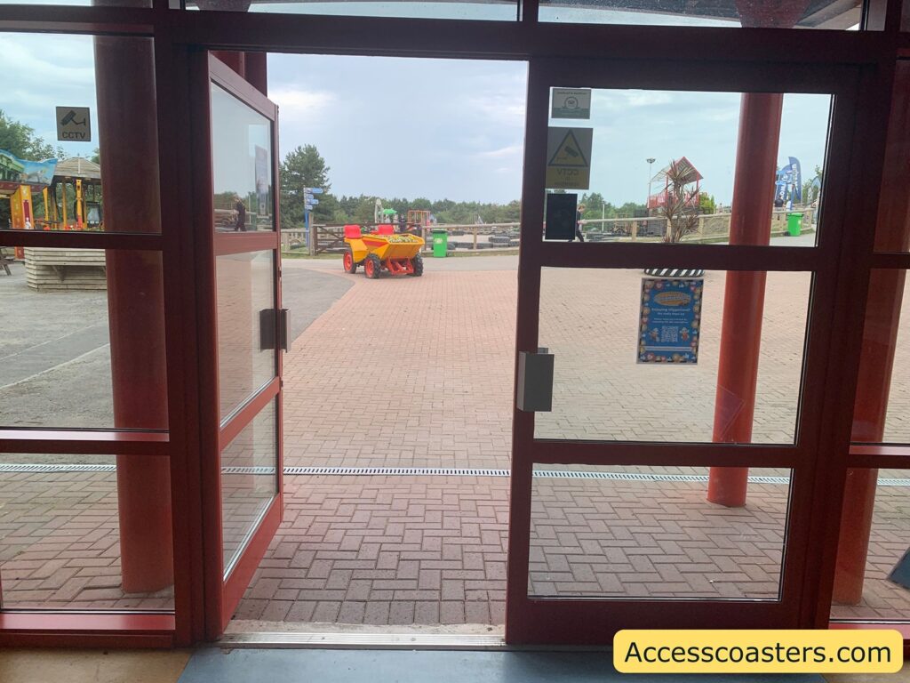 Glass entrance doors with a view outside to a paved courtyard. Just beyond the doors, a bright red and yellow children's ride-on digger or construction vehicle is parked.