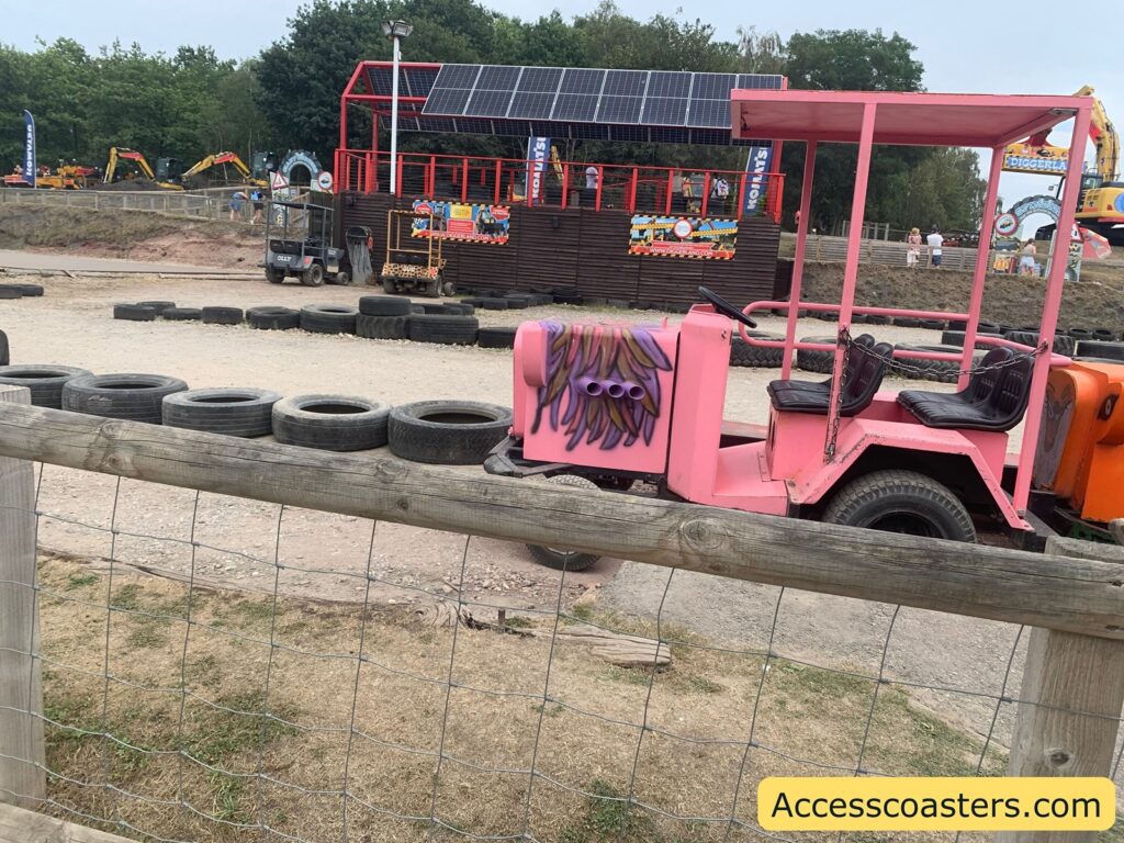 Pink vehicle shaped like a small truck or jeep, parked on a gravel surface within a fenced area. It has two seats, black steering wheel, and a painted engine block with purple pipes and flame designs on one side. Behind it, there’s an orange vehicle. The ground is surrounded by neatly lined tires, used as barriers. In the background,