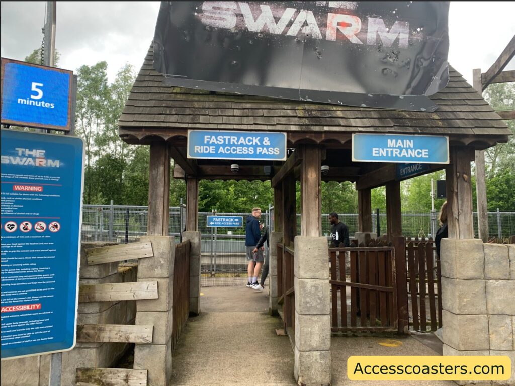 Entrance gate area for a theme park ride called "The Swarm." There are two entrance lanes: one labeled “Fastrack & Ride Access Pass” to the left, and one labeled “Main Entrance” to the right.