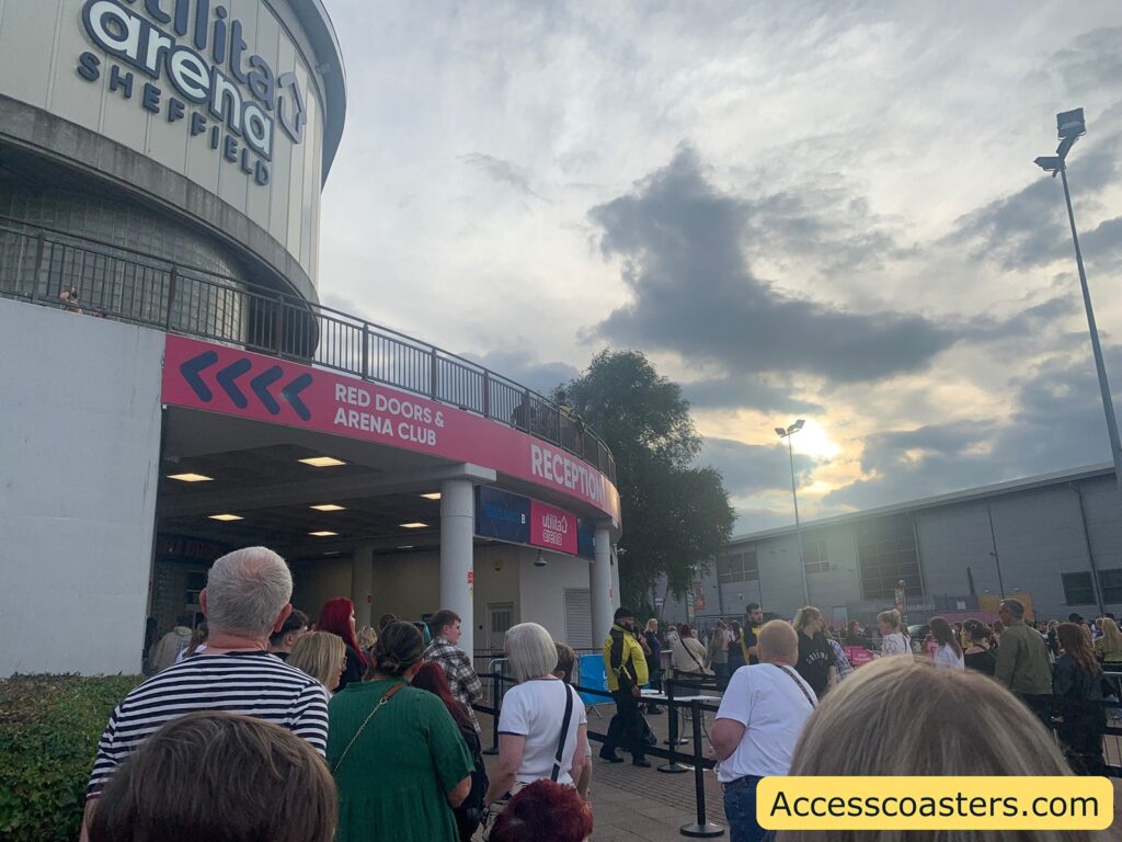Crowd of people queuing outside the entrance of Utilita Arena Sheffield. The arena’s large curved building dominates the left of the image, with its name and location displayed in bold letters. There is a pink sign pointing towards "Red Doors & Arena Club" and "Reception" above the entrance.