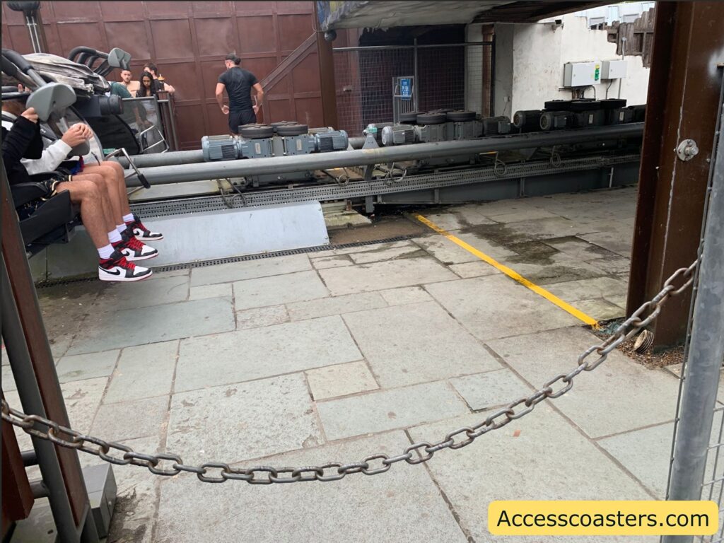Partially visible roller coaster loading platform with riders, focusing on two people seated in the front row of a ride vehicle with their legs dangling.