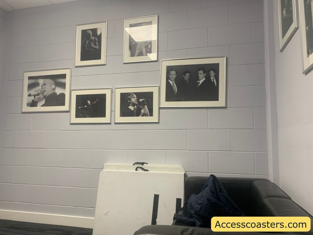 A wall with six black and white photographs in frames, showing musicians and a group of four men in suits. There is a small folding table leaning against the wall, a dark cushioned couch