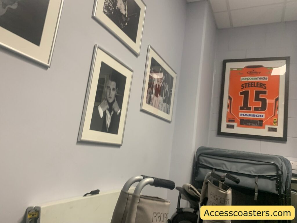photographs on a pale wall and a framed orange sports jersey labeled "STEELERS 15" with several sponsor logos including "purposemedia" and "HARSCO." Below the jersey, there are folded mobility aids such as wheelchairs.