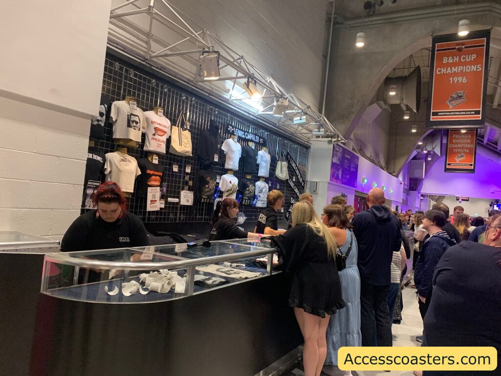 Merchandise stand at an indoor event venue. People are lined up at the counter, waiting to purchase various items which include t-shirts, hoodies, tote bags, and hats displayed on a grid wall behind the counter. Each item is labeled with a visible price tag in pounds. The merchandise primarily uses black, white, and orange colors and features text and graphic designs. Staff members in black shirts are assisting customers at the counter, which has some small items like wristbands inside the display case. Tote bag: £10 - T-shirt (with person’s face and “Problem” text): £20 - Long sleeve black shirt (right of tote bag): £30 - Orange cap: £20