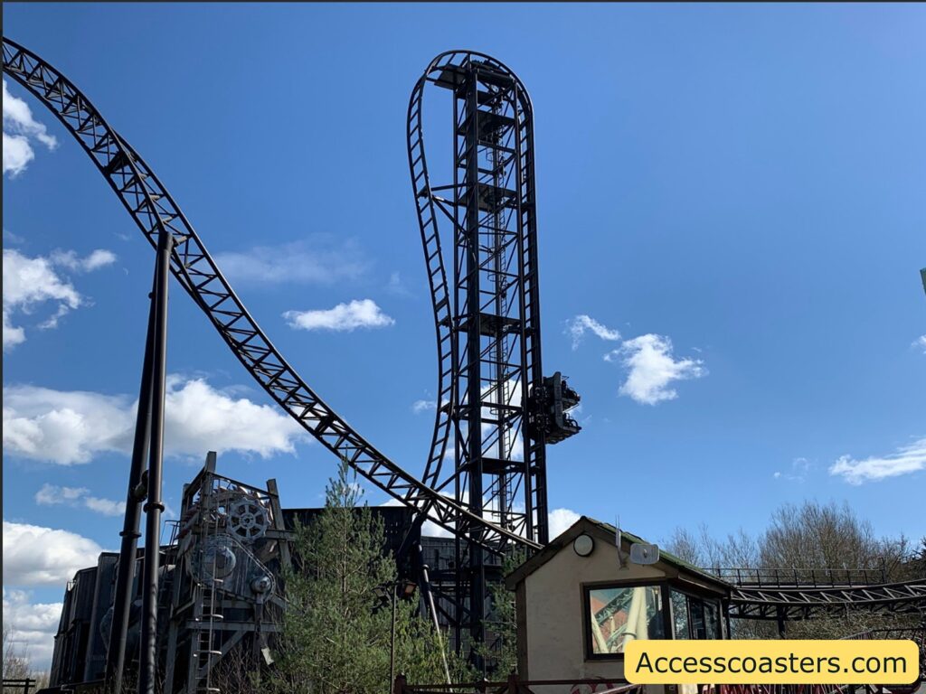 a prominent vertical track element, where the coaster track goes straight upward and then curves sharply at the top, forming a dramatic, twisting drop. There is a small building at the bottom right with a reflective window