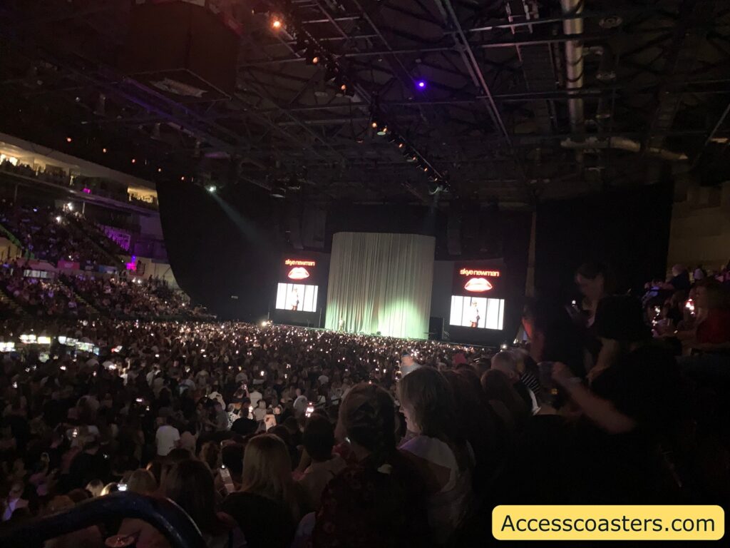 Indoor arena filled with a large audience, all facing a stage with a closed, illuminated curtain at the center. Two large screens on either side of the stage display the name “skyenewman” above a red graphic, each showing a performer or presenter. Many people in the crowd are holding up their phones with lights on