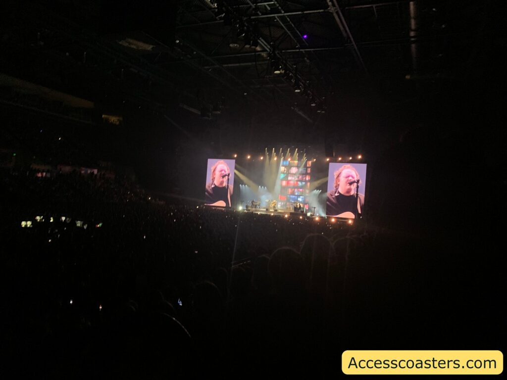 Concert scene inside a large indoor arena. The stage is brightly lit, with two tall screens on either side showing a close-up of a performer singing into a microphone and playing guitar. The crowd is dark, with many people visible