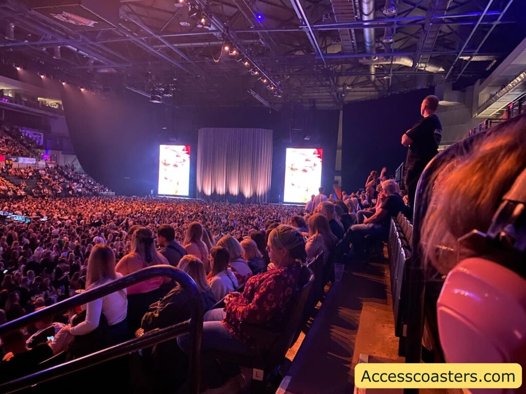 Be My AI Crowded indoor concert arena filled with people sitting and standing, waiting for the show to start. The stage has two large digital screens showing colorful images, with a large curtain hanging between them, concealing the stage setup. The arena is brightly lit with purple and blue lighting,