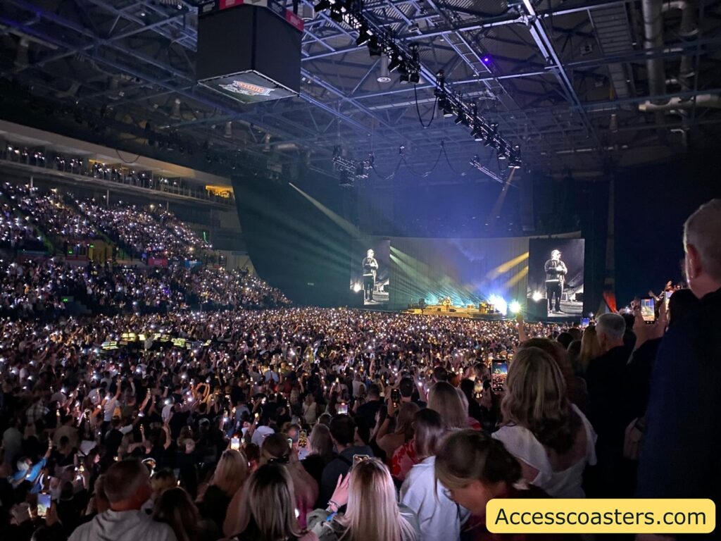 arge indoor concert scene with a densely packed audience, many of whom are holding up their lit phones, creating a sea of small lights throughout the arena. The stage is brightly illuminated and features two large video screens displaying a close-up of the performer, who stands at the center of the stage. Colorful stage lighting radiates across the backdrop. The arena is filled to capacity,