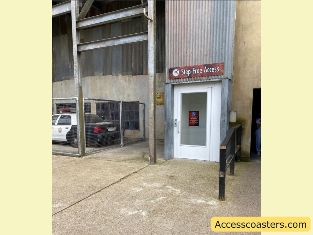 Step-free accessible entrance. The image shows a small lift or elevator with a glass door, designated for step-free access as indicated by a sign above the door that reads "Step-Free Access" along with the international symbol of accessibility (wheelchair symbol). There is another accessibility sign on the elevator door. The area surrounding the lift is paved with concrete. To the left of the lift, there's a fenced area containing a police car.