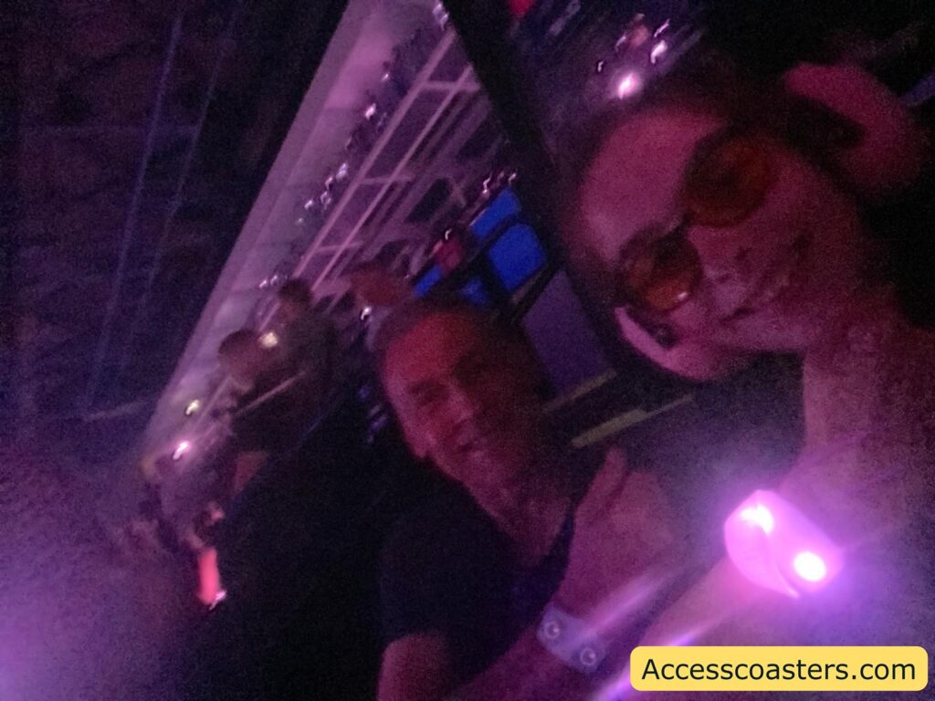 Dimly lit selfie of two people sitting indoors, possibly at a concert or an event. The lighting is tinted purple, and both individuals are smiling.