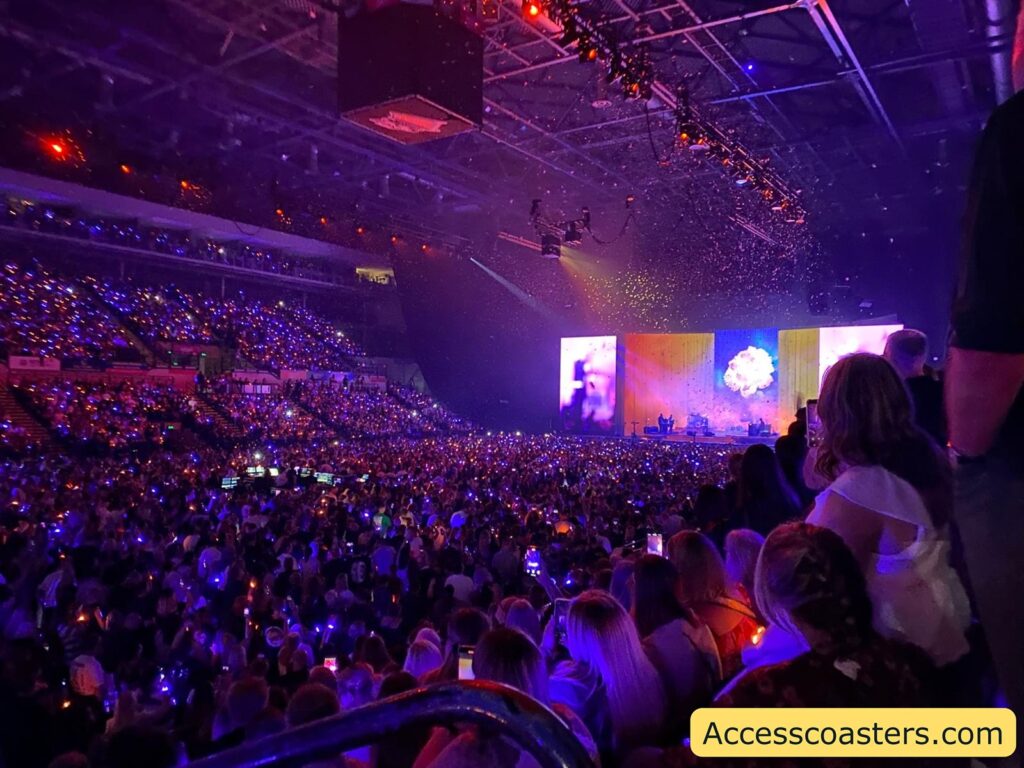 Packed indoor concert arena with thousands of people, all facing a brightly lit stage at the far end. The audience is bathed in purple and blue light, likely from LED wristbands or lights many are holding, creating a twinkling effect throughout the venue. On stage, multiple large screens display a colorful, abstract image with dominant yellow and blue hues, and there are visible performers and equipment. Confetti or glitter appears to be falling from the ceiling,
