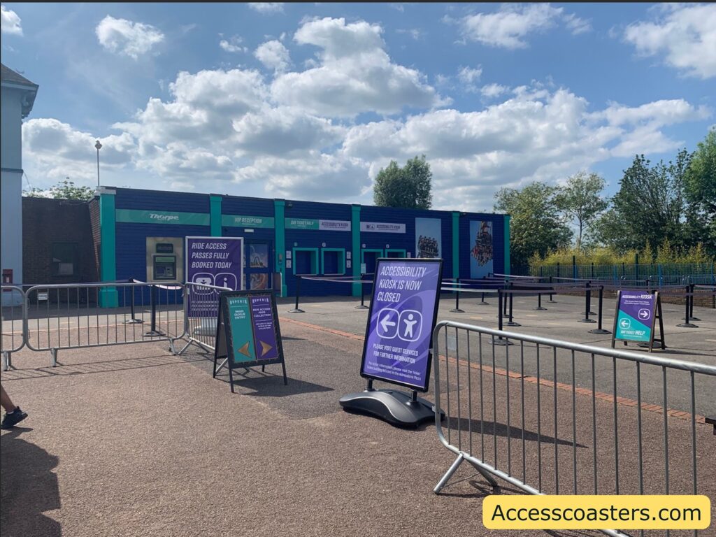 Outdoor theme park area with blue buildings and signs in front. Metal barriers create designated walkways. Several large purple signs are visible, one reading "RIDE ACCESS PASSES FULLY BOOKED TODAY" and another saying "ACCESSIBILITY KIOSK IS NOW CLOSED," with arrows for directions and advice to visit Guest Services for information.