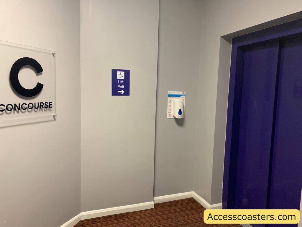 Elevator area with light grey walls, a purple elevator door on the right, a hand sanitizer dispenser mounted on the wall, and a purple sign indicating "Lift Exit" with a wheelchair symbol and a right-pointing arrow. There is also a partial sign on the left with the large letter "C" and "CONCOURSE"