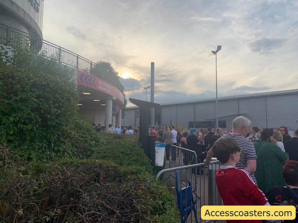 A line of people, including adults and children, waits outside a building with a sign reading "RED DOORS & ARENA CLUB" above the entrance. Metal barriers help organize the queue,