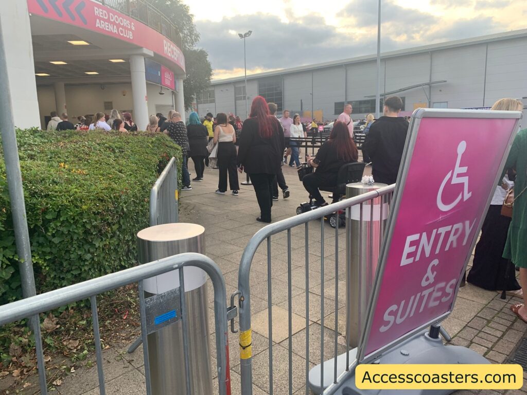 Crowd of people waiting outside an event venue. The scene shows people standing near the entrance labeled "RED DOORS & ARENA CLUB," under a covered area with directional pink signage. In the foreground, a large pink sign with a wheelchair symbol reads "ENTRY & SUITES," indicating accessible entry for disabled guests. Barricades help guide the crowd,
