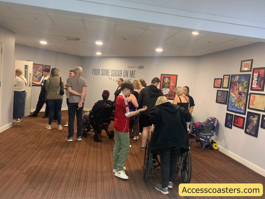 Crowd of people, some using wheelchairs, in a hallway with music-themed wall decorations and framed posters. The wall features a large text that reads "POUR SOME SUGAR ON ME" with smaller text underneath. Some people are standing while others are sitting or assisting those in wheelchairs.