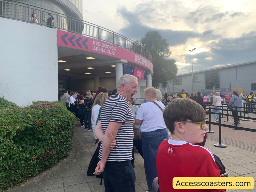 Queue of people outside an arena entrance, with some standing near a sign that reads “RED DOORS & ARENA CLUB RECEPTION.” The sign is on a pink banner above a doorway. There are black metal barriers forming the queue area, and more people are visible outside and inside the entrance area.