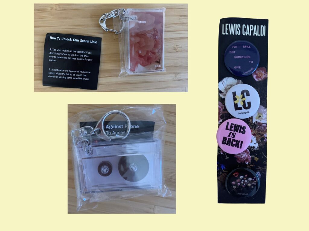 Top left: A clear keychain with pink fragments inside, next to a black card titled "How To Unlock Your Secret Link!" with instructions for using your mobile phone to unlock a secret link by tapping it on the included cassette item.

Bottom left: A clear plastic keychain shaped like a cassette tape inside a plastic bag, accompanied by an instruction sheet partially visible behind it.

Right side: A black card with "LEWIS CAPALDI" printed at the top, displaying four pin-back buttons. From top to bottom: a purple button with white text reading "I'VE STILL GOT SOMETHING TO GIVE", a white button with abstract "LC" initials, a pink button with "LEWIS IS BACK!", and a black button featuring a floral pattern. The black card itself has a floral design in the background.