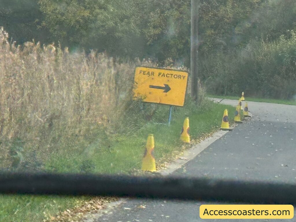 Yellow road sign reading “FEAR FACTORY” with an arrow, positioned beside a narrow driveway and lined with small cones, directing cars towards the event parking area.
