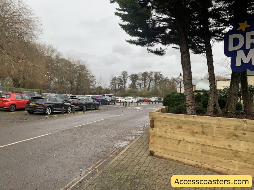 Hardstanding disabled parking bays at Drayton Manor, showing a row of cars parked alongside a wide road with a wooden planter and trees near the entrance.
