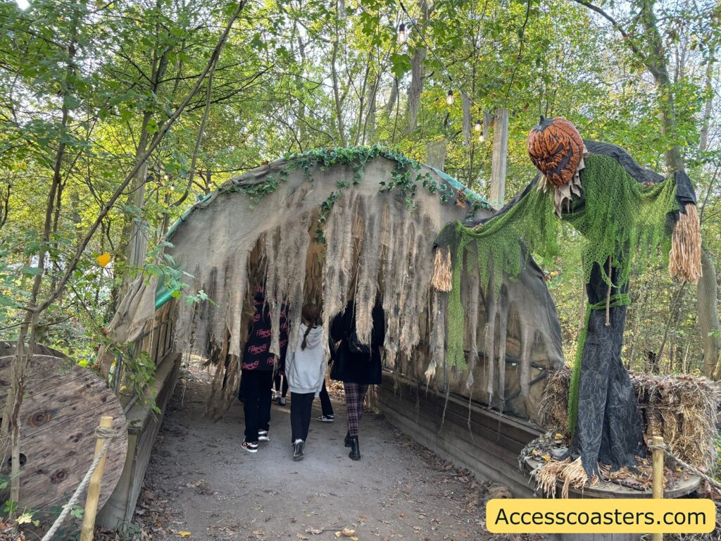 people walk into a scare zone tunnel made of tattered fabric and netting, with a tall, menacing scarecrow figure standing to the right of the entrance.